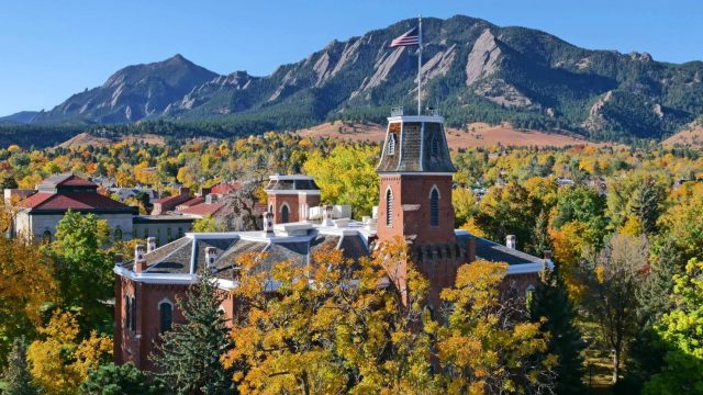 Aerial view of CU's Old Main spire and roof poking above trees in full yellow and orange fall colors. Flatirons can be seen in background against a clear blue sky.