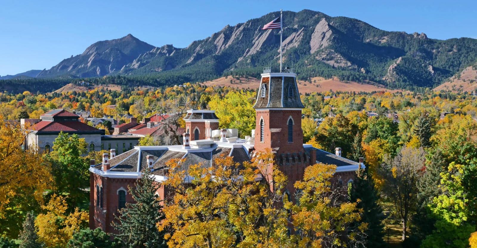 Aerial view of CU's Old Main spire and roof poking above trees in full yellow and orange fall colors. Flatirons can be seen in background against a clear blue sky.
