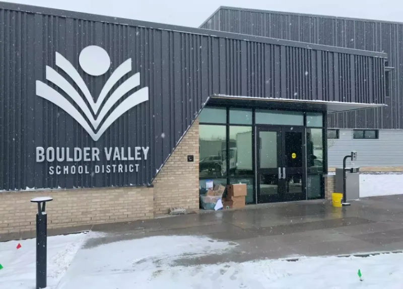 Exterior via of the Boulder Valley School District main entrance showing large logo on building to the left of glass doors with snow on the ground.