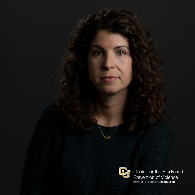 Headshot of a white woman with dark brown curly hair wearing a dark green sweater.