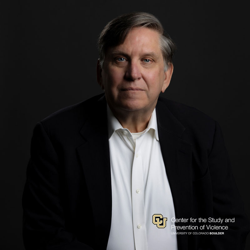 Headshot of white man with short grey hair wearing a white button down shirt and a black blazer.
