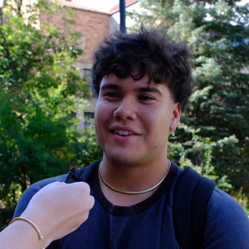 Person with short dark brown hair standing in front of trees and wearing a navy shirt and backpack.