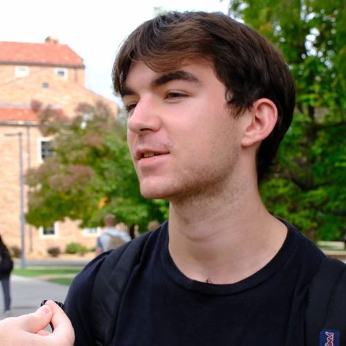 Person with short brown hair and black shirt standing in front of trees and buildings.