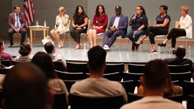 A group of 8 panelists sit in chairs on a stage in front of an audience: 3 Latina women, 2 white women, 1 Black man, and 1 white male moderator.