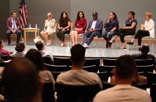 A group of 8 panelists sit in chairs on a stage in front of an audience: 3 Latina women, 2 white women, 1 Black man, and 1 white male moderator.