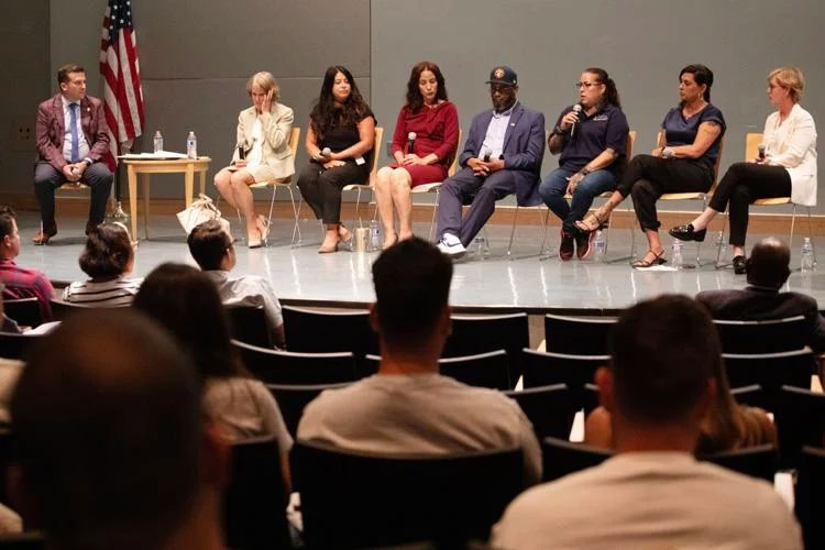 A group of 8 panelists sit in chairs on a stage in front of an audience: 3 Latina women, 2 white women, 1 Black man, and 1 white male moderator.