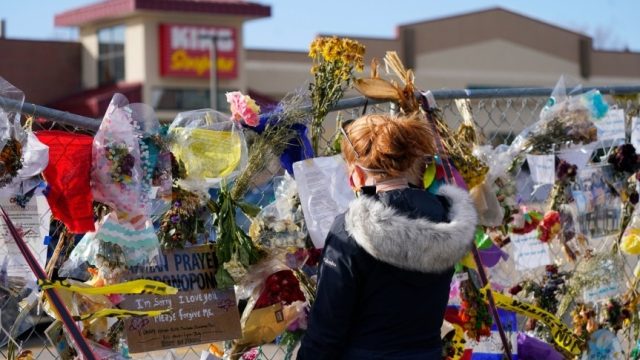 A woman with red hair reads a piece of paper left on the fence barricade in front of King Soopers. The fence is covered with flower bouquets and notes.