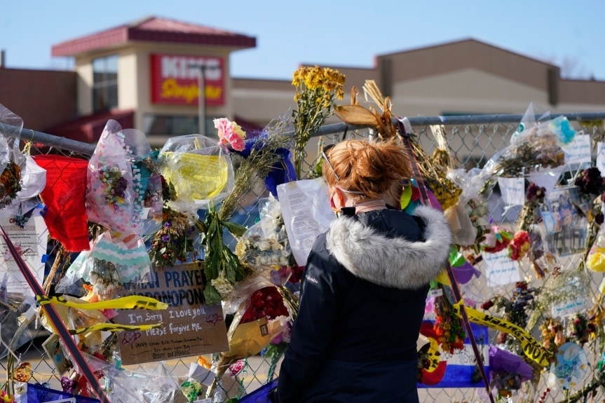 A woman with red hair reads a piece of paper left on the fence barricade in front of King Soopers. The fence is covered with flower bouquets and notes.