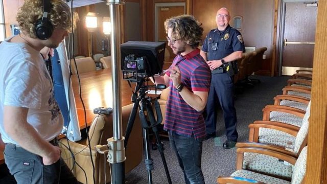 Two white men with curly hair wearing headphones look at video equipment and lights set up in a conference room with large table and several chairs. White male CUPD deputy chief stands in background in uniform.