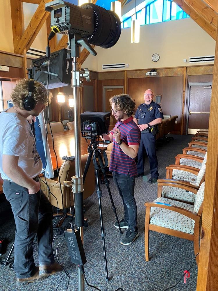 Two white men with curly hair wearing headphones look at video equipment and lights set up in a conference room with large table and several chairs. White male CUPD deputy chief stands in background in uniform.