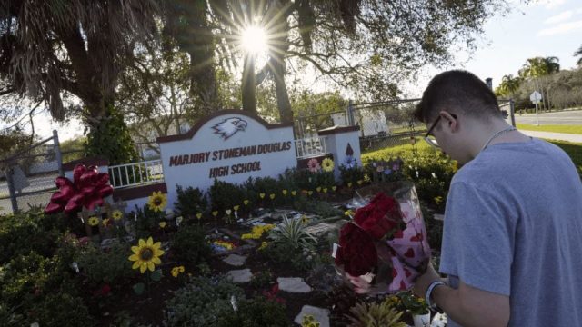 A man with very short hair and glasses stands holding two bouquets of roses in front of the Marjory Stoneman Douglas High School sign in front of which is a beautiful garden with perennials and stone steps, balloons, and decorative metal yellow flowers.