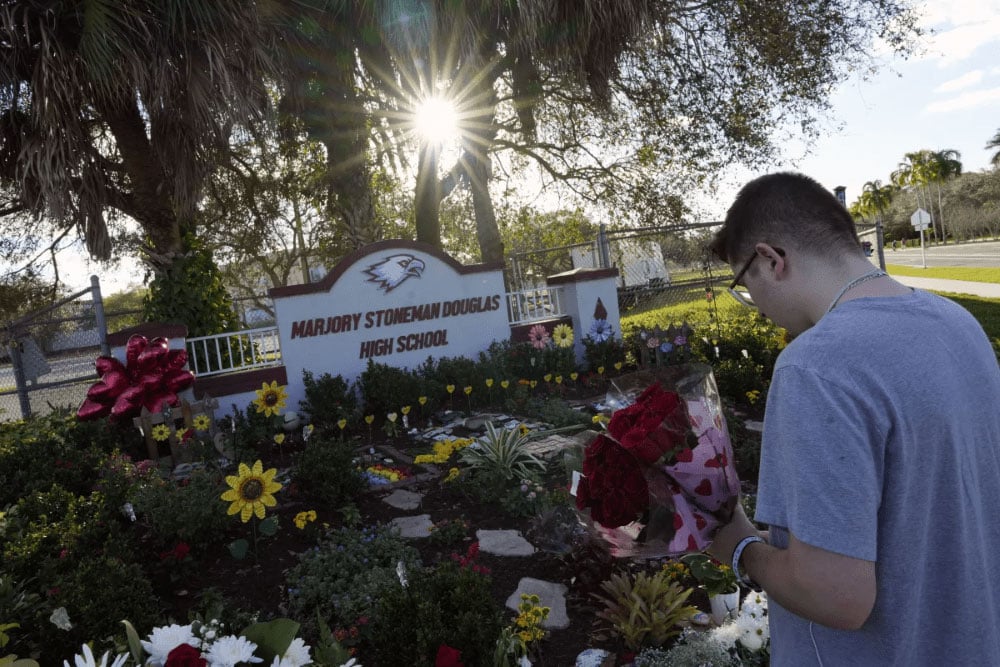 A man with very short hair and glasses stands holding two bouquets of roses in front of the Marjory Stoneman Douglas High School sign in front of which is a beautiful garden with perennials and stone steps, balloons, and decorative metal yellow flowers.