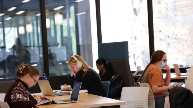 Three white and one dark skinned female students sit at tables looking at their laptops. One is wearing headphones and another is wearing a medical mask.