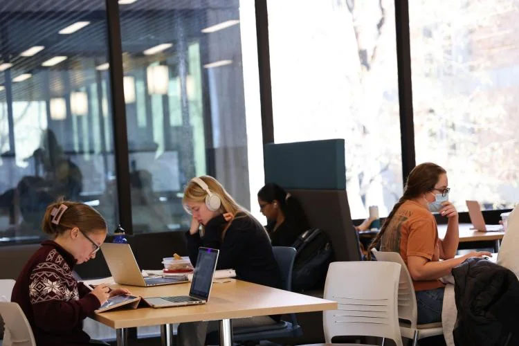 Three white and one dark skinned female students sit at tables looking at their laptops. One is wearing headphones and another is wearing a medical mask.