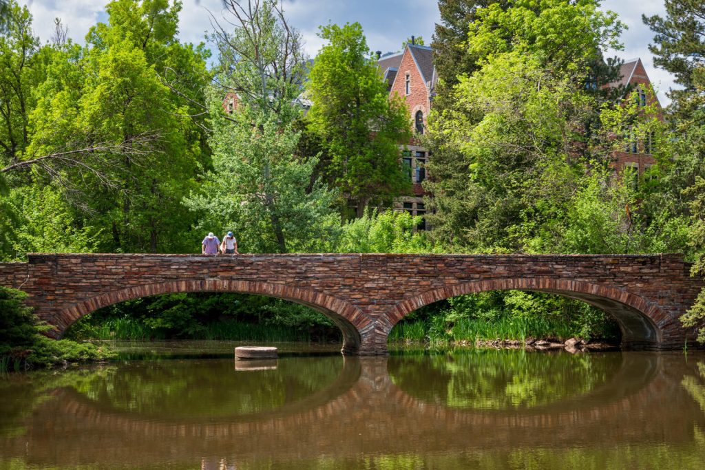 Two people stand on the double-arched, stone bridge overlooking Varsity Lake. Green trees and peak of campus building can be seen in the background. (Photo by Glenn J. Asakawa/University of Colorado)