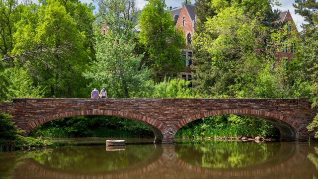 Two people stand on the double-arched, stone bridge overlooking Varsity Lake. Green trees and peak of campus building can be seen in the background. (Photo by Glenn J. Asakawa/University of Colorado)